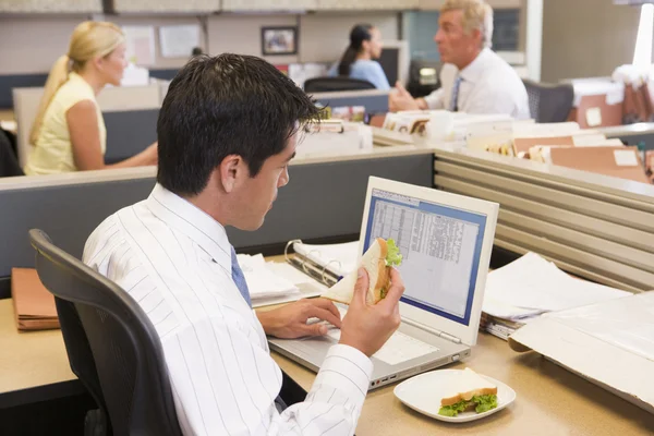 Businessman in cubicle at laptop eating sandwich