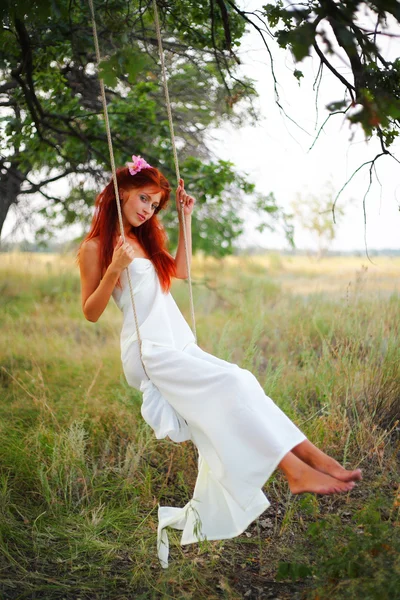 The red-haired girl in a white dress shakes on a swing in a grove
