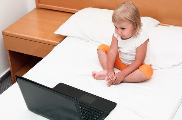 Ein kleines Kind sitzt mit einem Laptop auf dem Bett und schaut auf den Bildschirm — Stockfoto