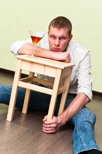 П'яний чоловік сидить на підлозі зі склянкою алкоголю і тримає табурет — стокове фото