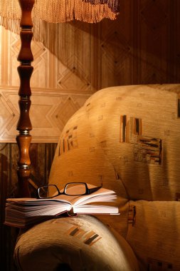 Home interior with luminous floor lamp near armchair. Open book and spectacles lying on armchair clipart