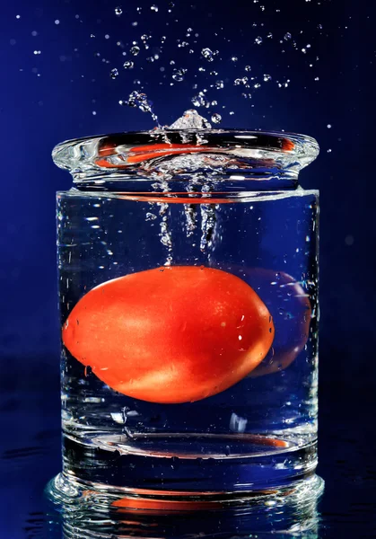Красный помидор падает в стекло с водой на глубокой синей — стоковое фото
