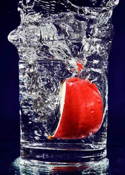 Bit av rött äpple faller ner i glas med vatten på djup blå — Stockfoto