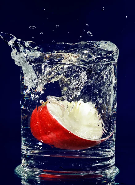 Bit av rött äpple faller ner i glas med vatten på djup blå — Stockfoto