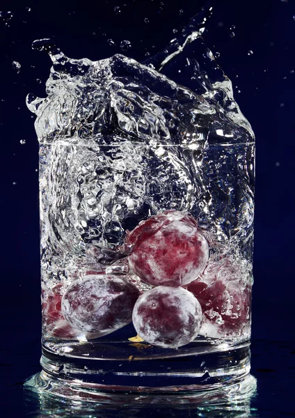 Ramo de uvas rojas cayendo en vaso con agua en blu profundo — Foto de Stock