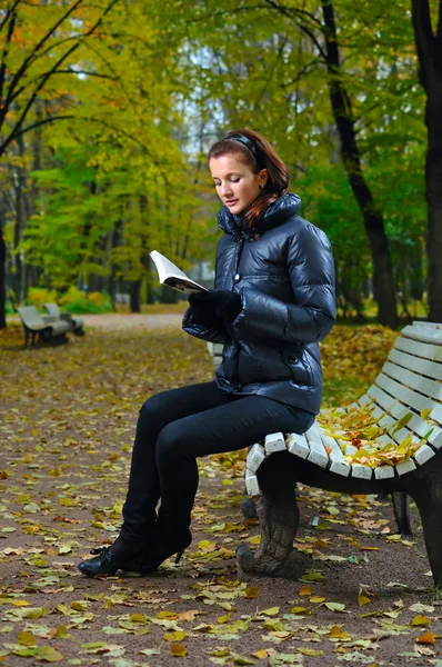 Jovem mulher está sentada em um banco em um parque de outono e leitura — Fotografia de Stock