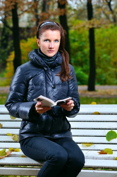 Genç kadın bir sonbahar park ve okuma bir bankta oturuyor — Stok fotoğraf