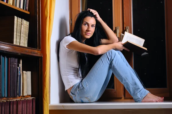 Hermosa mujer está sentada en un alféizar de la ventana y sosteniendo un libro — Foto de Stock