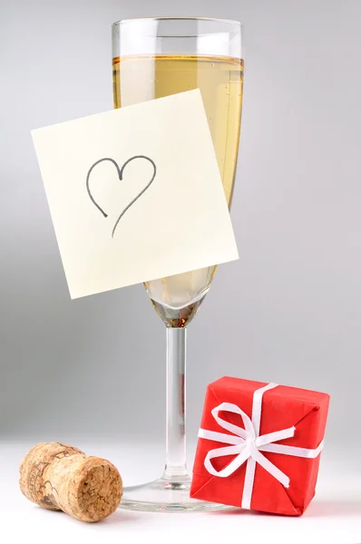 Champagne glass with heart on sticky note, red gift box and cork — Stock Photo, Image