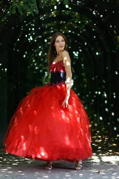 Schöne Frau in rotem Kleid, die in einer Gasse im Park spaziert — Stockfoto