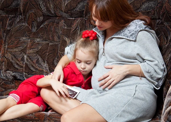 Raskaana oleva nainen tyttärensä kanssa katsoo kuvaa sohvalla — kuvapankkivalokuva