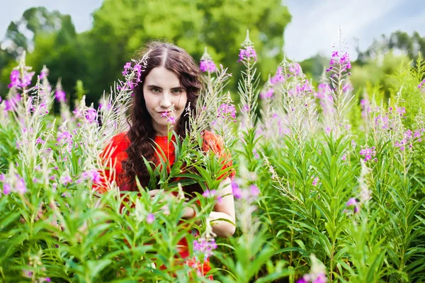 Chica sonriente en vestido rojo mirando a través de las flores en el mea —  Fotos de Stock