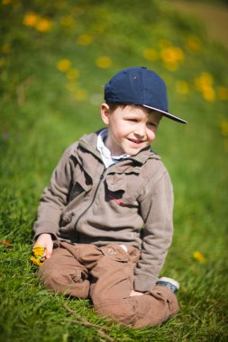 Cute 4 years old boy with dandelion sitting on grass clipart