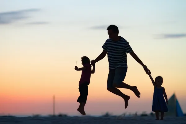 父亲和他两个孩子跳跃和日落时分在海滩上玩的 Silhouettes — 图库照片