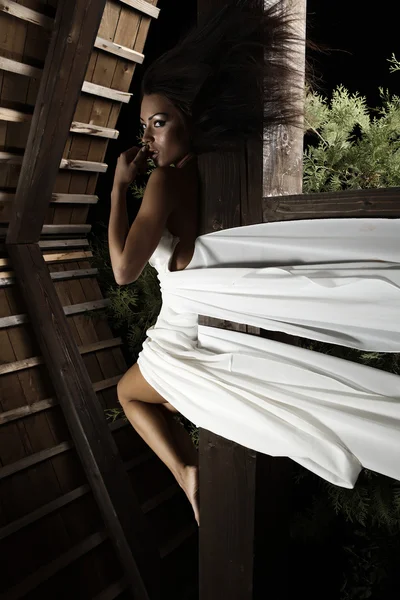 Atraktivní opálený dívky v bílých šatech představuje na dřevěný hranol. — Stock fotografie