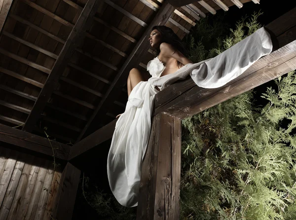 Attractive suntanned girl in white dress poses on a wooden beam. — Stock Photo, Image