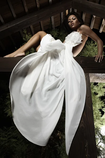 Attraktives, sonnengebräuntes Mädchen im weißen Kleid posiert auf einem Holzbalken. — Stockfoto