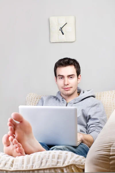 笑的年轻人使用的便携式计算机和躺在家里的沙发上 — 图库照片