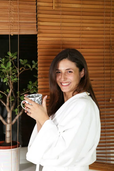 Avslappnad kvinna i morgonrock hemma med te eller kaffe Stockbild