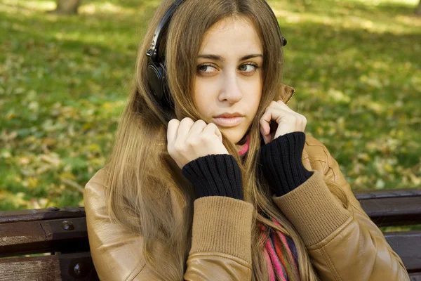 年轻白种女人用耳机在秋天公园坐在长凳上 周围很多多彩叶面肥秋 — 图库照片