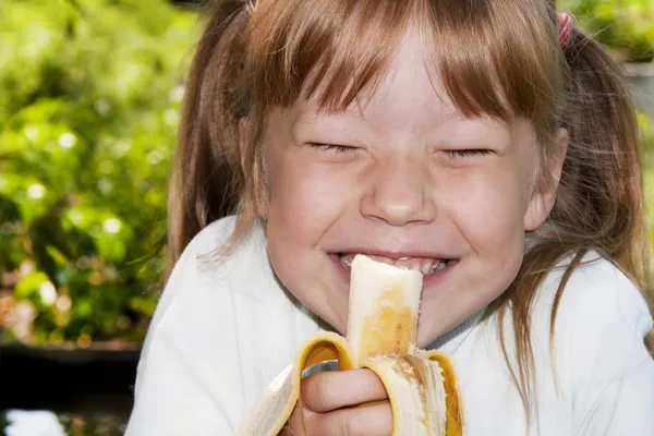 Little girl in the outdoors eats a banana and laughs — Stock Photo, Image