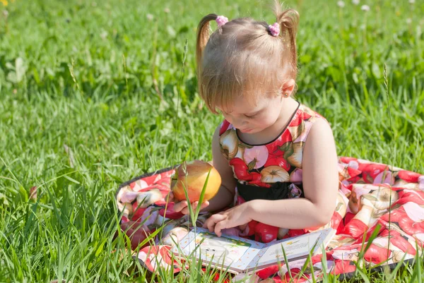 Flickan äta ett äpple och ser boken på gräset — Stockfoto