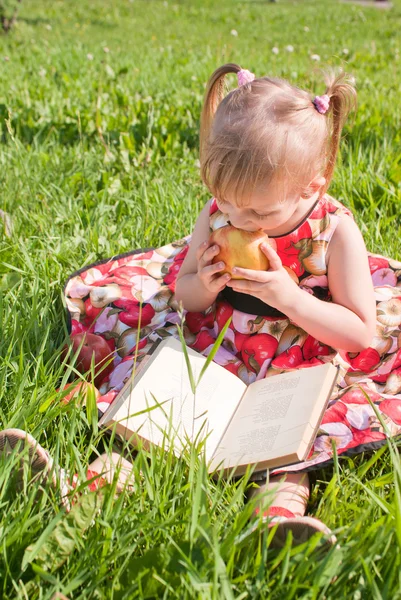 Girl eating an apple and looking book on the grass — Stock Photo, Image
