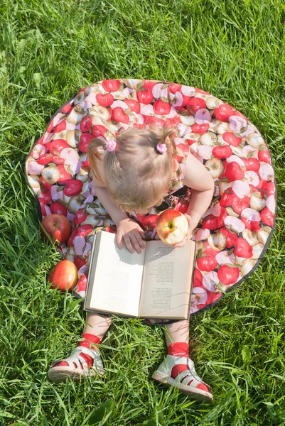 Κορίτσι τρώει ένα μήλο και κοιτάζοντας το βιβλίο στο γρασίδι — Φωτογραφία Αρχείου