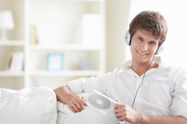 Attractive man with a disk in his hands listening to music through headphones on the sofa clipart