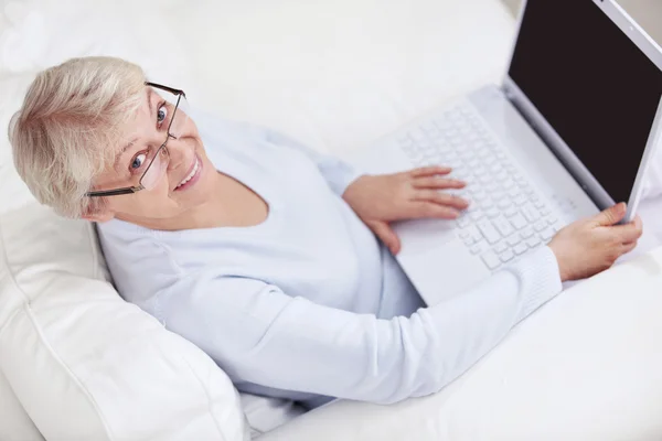 一名老妇在白色沙发上的一台笔记本电脑 — 图库照片