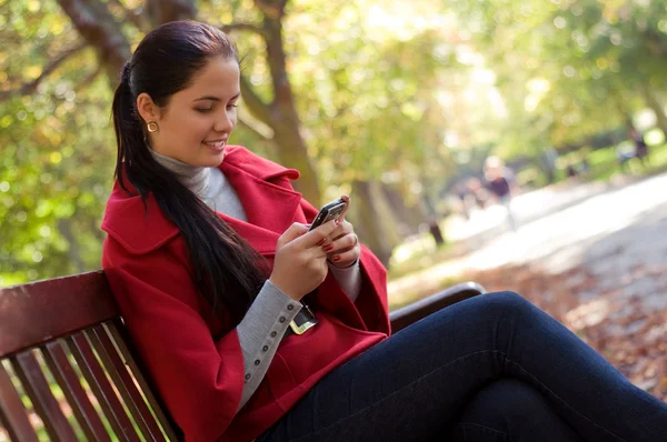 Unga kaukasiska kvinna med en mobiltelefon, sitter i en park på ett — Stockfoto