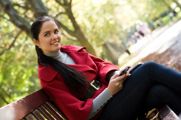 Sms を読んで公園木製のベンチで座っている携帯電話を持つ若い白人女性 — ストック写真