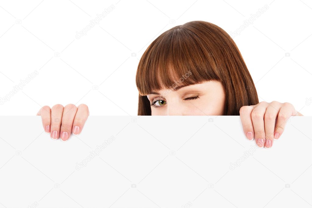 Winking young woman peeking over blank billboard sign isolated on white