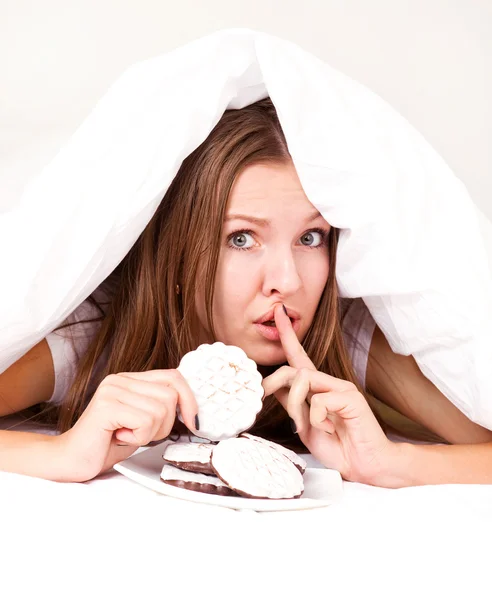 美しい若い女性は毛布の下に隠れているとベッドの中でクッキーを食べて — ストック写真