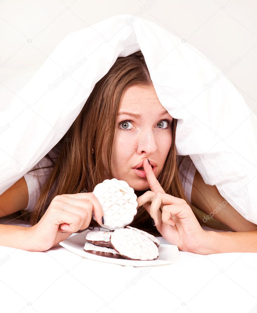 Beautiful young woman hiding under the blanket and eating cookies in bed