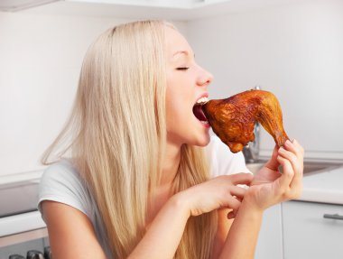 Beautiful young blond woman eating chicken in the kitchen at home clipart