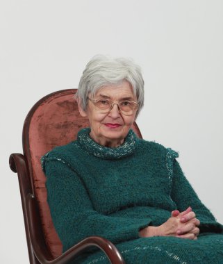 Portrait of an old woman sitting on a wooden rocking chair with fingers crossed, against a grey background. clipart