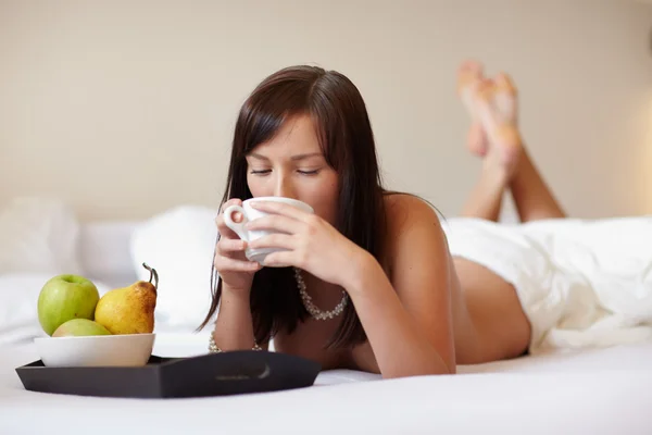 Молода красива жінка п'є каву, лежачи — стокове фото