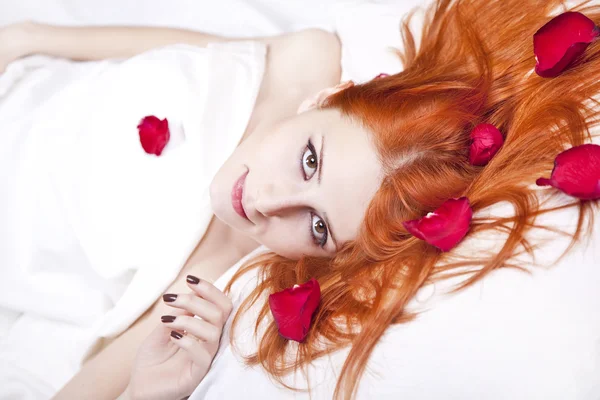 バラの花びらとベッドの中で美しい赤い髪の少女. — ストック写真