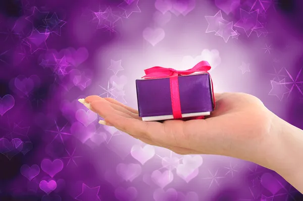 Vrouwelijke hand met paarse geschenk op sterrenhemel hart achtergrond — Stockfoto