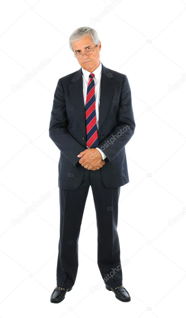 Serious middle aged businessman in a suit and tie standing with hands clasped in front peering over the top of his glasses. Full length over a white background.