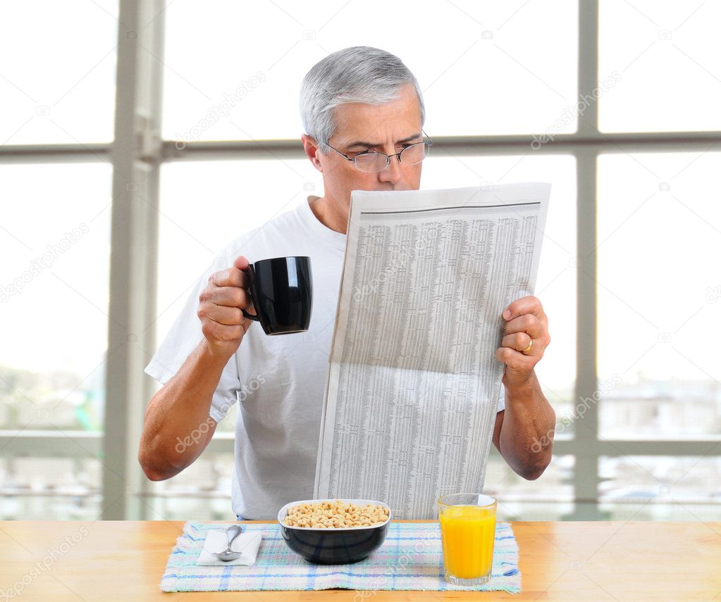 Casually dressed middle aged man reading the morning newspaper in front of large window with breakfast cereal, juice and coffee in front of him. Square format.