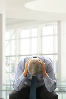 Seated Middle Aged Businessman Doubled Over with His Head in Hands in a modern business office. Vertical Format. clipart
