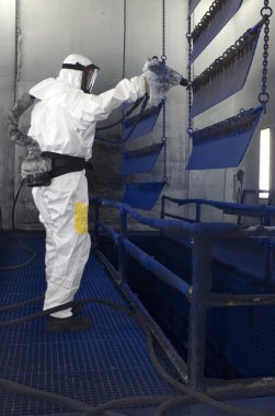 Man, wearing a gas mask and a paper protective suit with respiration aid spray painting semi finished metal parts in a factory clipart