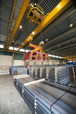 Huge overhead crane, handling pallets containing steel sheet at a warehouse clipart