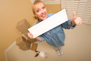 Excited Woman with Thumbs Up and Doggy Holding Blank Sign Near Moving Boxes in Empty Room Taken with Extreme Wide Angle Lens. clipart