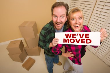 Goofy Couple Holding We've Moved Sign in Room with Packed Cardboard Boxes. clipart