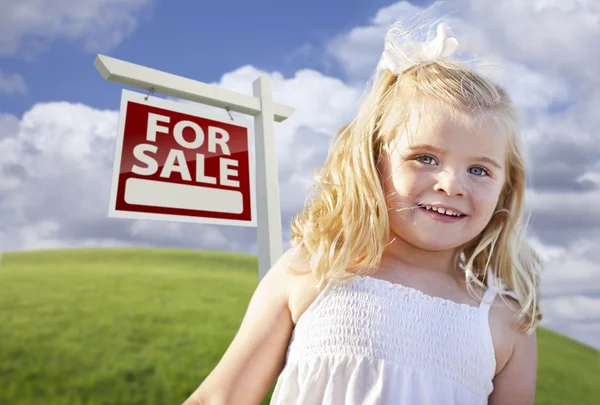 Улыбающаяся симпатичная девушка в поле с вывеской на продажу недвижимости — стоковое фото
