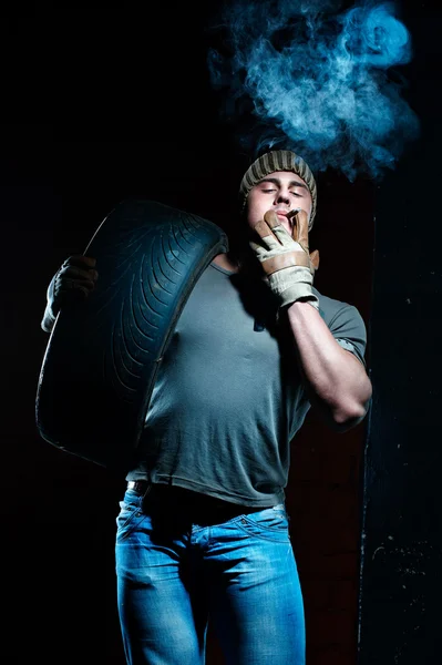 Jovem homem muscular com pneu fumando — Fotografia de Stock