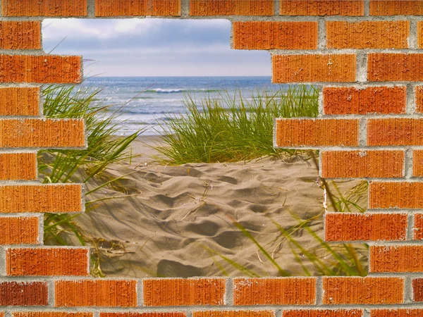 Caminho arenoso com grama de praia atrás de um buraco em uma parede de tijolo — Fotografia de Stock